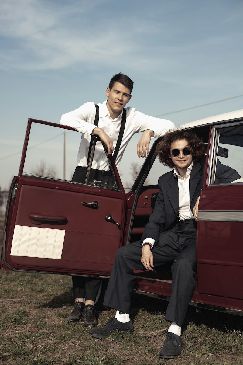 Two sharply dressed young men posing with a vintage maroon car